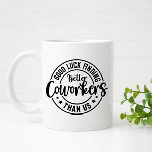 May include: White ceramic mug with a black circular design that reads "Good Luck Finding Better Coworkers Than Us." The mug has a curved handle and is set against a white background with a small green plant.