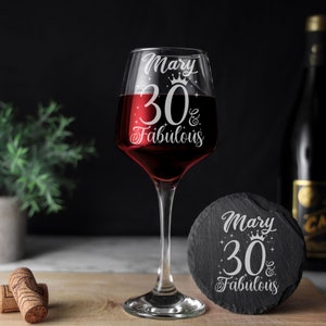 May include: A clear glass wine glass filled with red wine and a round black slate coaster, both featuring white etched text that reads Mary 30 & Fabulous with a crown and star accents. Two corks are positioned on the left side of the wooden surface.