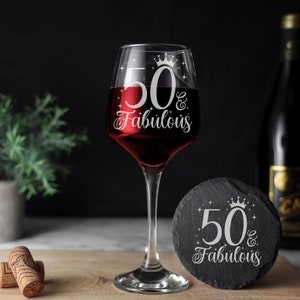 May include: A clear wine glass with a black stem and base. The glass is engraved with the text "50 & Fabulous" and a crown. A black slate coaster with the same text and a crown is next to the glass.