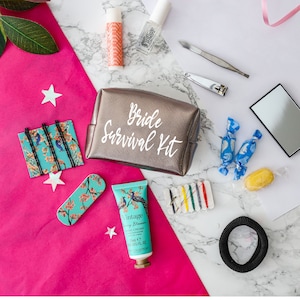 May include: A silver cosmetic bag with the text "Bride Survival Kit" on it. The bag is surrounded by various items, including hair ties, bobby pins, nail clippers, hand cream, and sweets.