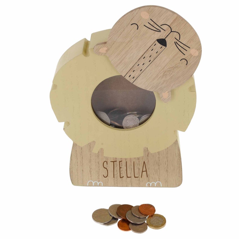 May include: A wooden lion money box with a slot on the back and a removable lid. The money box is painted yellow and has a brown lion face with a black nose and whiskers. The words "STELLA" are printed on the front of the money box.