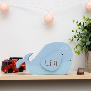 May include: A light blue wooden whale money box with a slot on the back for coins. The whale has a smiling face and small hearts painted on its back. The name "LEO" is written on the whale's side in brown paint.