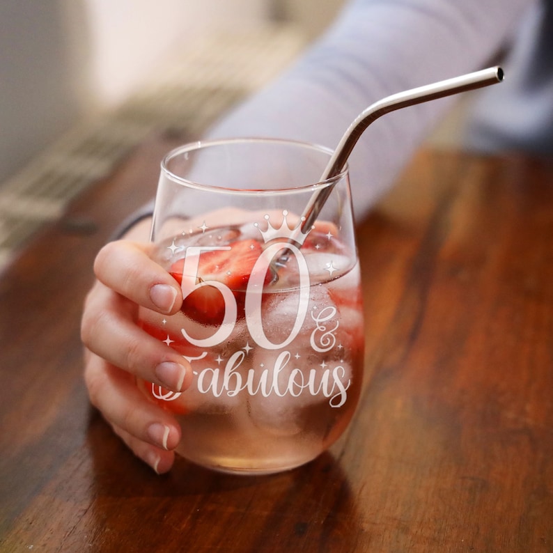 May include: A clear glass with a straw and ice, filled with a pink drink and strawberries. The glass has the text "50 & Fabulous" etched on it.
