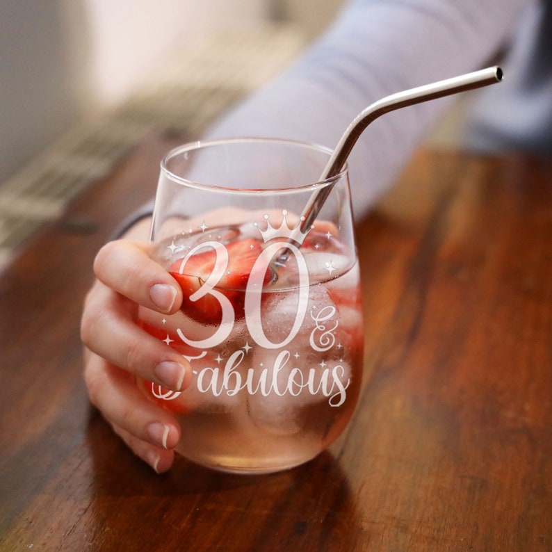 May include: A clear glass wine glass with a straw and ice cubes. The glass has the text "30 & fabulous" etched on it. There are strawberries in the glass.