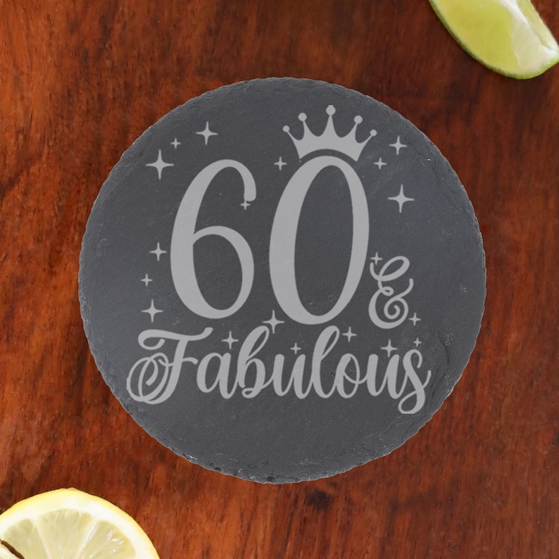 May include: A round black slate coaster with a silver inscription that reads "60 & Fabulous" with a crown above the number 60. The inscription is surrounded by small silver stars.