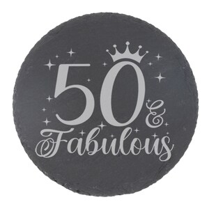 May include: A black slate round coaster with a silver inscription that reads "50 & Fabulous" with a crown above the number 50.