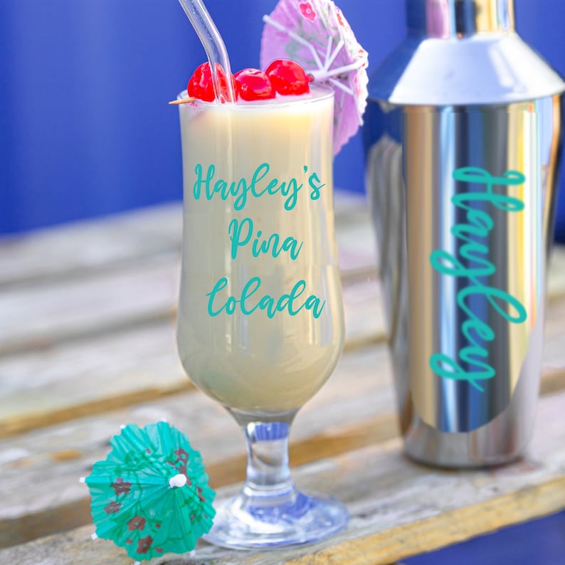 Puede incluir: Un vaso de Pina Colada de Hayley con una sombrilla de papel rosa y blanca y cerezas maraschino. La bebida est&aacute; en un vaso alto con tallo y base transparente. El vaso est&aacute; sobre una superficie de madera.