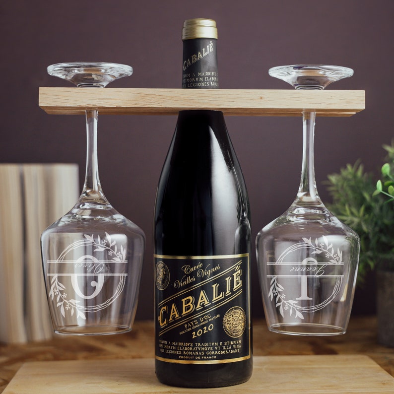 Puede incluir: Un soporte de madera para botellas de vino y vasos con dos vasos. La botella es una botella de vino tinto oscuro con una etiqueta dorada que dice "Cuv&eacute;e Vieilles Vignes CABALIE PAYS D'OC 2010". Los vasos son transparentes y tienen un dise&ntilde;o floral con una inicial grabada en cada uno.