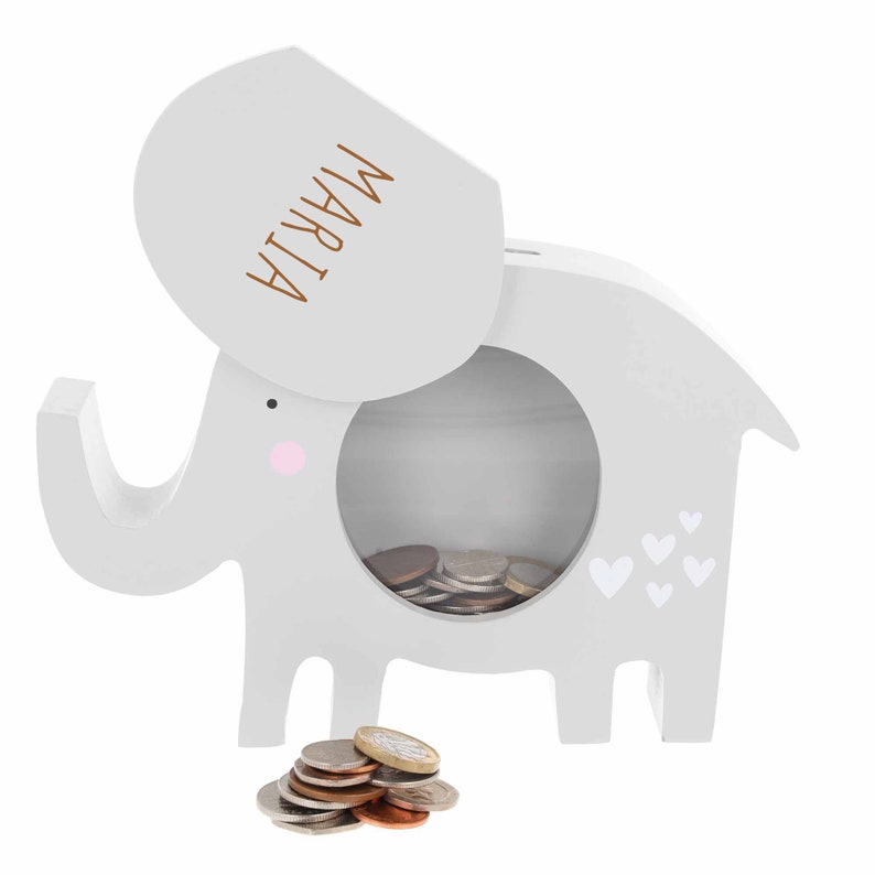 May include: Grey wooden elephant money box with a slot on the side and a removable lid. The lid has the name "MARIJA" written on it. The elephant has pink cheeks and white hearts on its side. There are coins in the money box and on the floor.