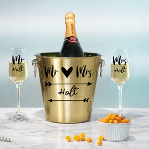 May include: Gold champagne bucket with two champagne flutes and a bottle of champagne. The bucket and flutes are decorated with the text "Mr. & Mrs. Holt" and arrows.
