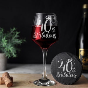 May include: A clear wine glass with red wine inside. The glass has a white design that says "40 & Fabulous" with a crown on top of the 40. A black slate coaster with the same design is next to the glass.