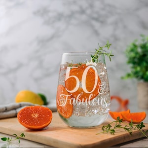 May include: A clear glass wine glass with the text "50 & Fabulous" etched on the side. The glass is filled with a clear liquid and orange slices.