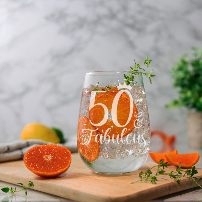 May include: A clear glass wine glass with the text "50 & Fabulous" etched on the side. The glass is filled with a clear liquid and orange slices.
