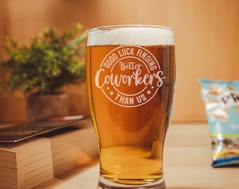 May include: A tall beer glass filled with a light amber liquid and topped with a foamy head. The glass has the words "Good Luck Finding Better Coworkers Than Us" etched on the front. The glass sits on a wooden surface.