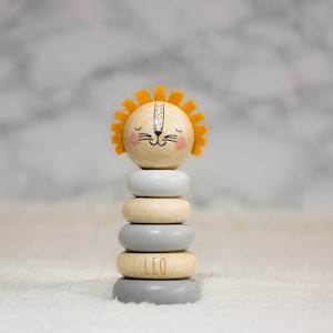 May include: A wooden stacking toy shaped like a lion with a yellow mane. The toy has five rings, alternating between grey and natural wood. The bottom ring has the name "LEO" inscribed on it.