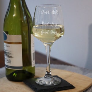 Peut inclure: Un verre à vin en verre transparent avec le texte "Don't Ask", "Bad Day" et "Good Day" gravé sur le côté. Le verre est rempli de vin blanc et repose sur un dessous-de-verre noir.