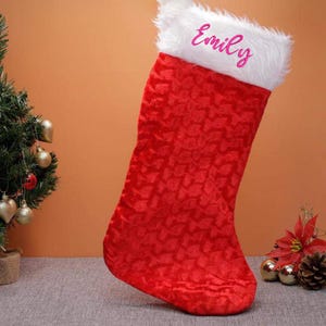 May include: Red and white Christmas stocking with a white faux fur cuff. The stocking has a snowflake pattern and the name "Emily" is embroidered on the cuff in pink.