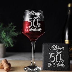 May include: A personalized wine glass with the text "Alison 50 & Fabulous" etched on it. The glass is filled with red wine and is sitting on a wooden surface. There is a black slate coaster with the same text on it.