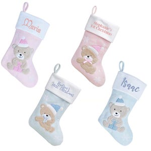 May include: Four personalised Christmas stockings featuring a teddy bear design. The stockings are in pink and blue with white trim and snowflake patterns. The names on the stockings are "Maria", "Stephanie's 1st Christmas", "Hudson's First Christmas", and "Isaac".