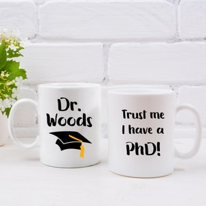 Peut inclure: Deux tasses en céramique blanche avec du texte noir. La tasse de gauche dit "Dr. Woods" avec un bonnet de graduation noir et un gland jaune. La tasse de droite dit "Faites-moi confiance, j'ai un doctorat !"