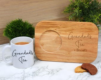 May include: A white mug with the text "Grandad's Tea" next to a wooden tea tray with a circular indentation and the words "Grandad's Tea + Biscuits". Two biscuits are on the marble surface.