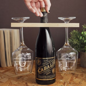 May include: A wooden wine rack with two wine glasses and a bottle of red wine. The glasses are engraved with the letters "O" and "T" and floral designs. The wine bottle label reads "Cuv&eacute;e Vieilles Vignes CABALIE PAYS DOC 2010".