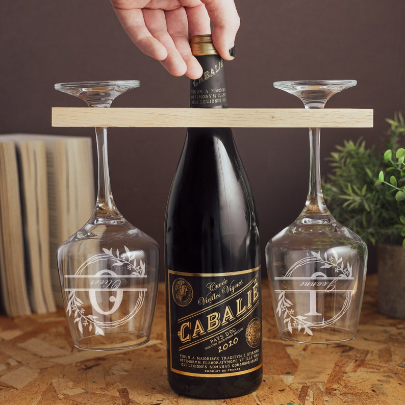 May include: A wooden wine rack with two wine glasses and a bottle of red wine. The glasses are engraved with the letters "O" and "T" and floral designs. The wine bottle label reads "Cuv&eacute;e Vieilles Vignes CABALIE PAYS DOC 2010".
