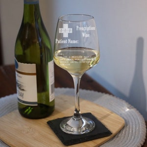 May include: A clear glass wine glass with a white design that says "Prescription Wine" and "Patient Name:" with a medical symbol. The glass is filled with white wine and is sitting on a black coaster.