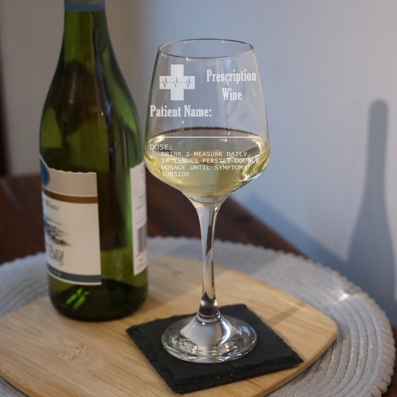 May include: A clear glass wine glass with a white design that says "Prescription Wine" and "Patient Name:" with a medical symbol. The glass is filled with white wine and is sitting on a black coaster.