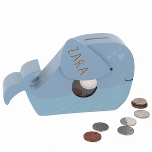 May include: A light blue wooden whale money box with a slot for coins. The whale has a white eye and a small mouth. The name "ZARA" is written on the side of the whale in brown lettering. There are several coins scattered on a white background.