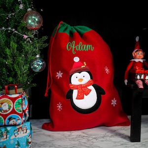 May include: Red Christmas gift bag with a penguin design and the name "Adam" embroidered on the front. The bag is made of felt and has a drawstring closure.