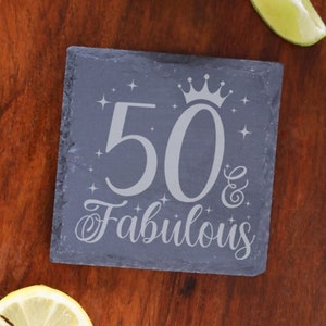 May include: A gray slate coaster with the text "50 & Fabulous" in white lettering. A crown is above the number 50. The coaster is decorated with small white stars.