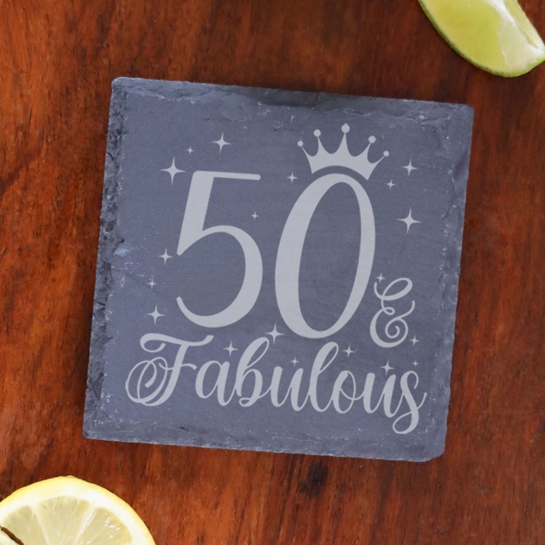May include: A gray slate coaster with the text "50 & Fabulous" in white lettering. A crown is above the number 50. The coaster is decorated with small white stars.