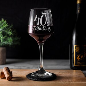 May include: A clear wine glass with a stem and a black coaster. The glass has a white design that says "40 & Fabulous" with a crown above the number 40.