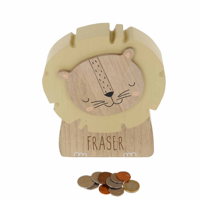 May include: A wooden lion money box with a yellow mane and a brown face. The money box has the name "FRASER" written on the front.