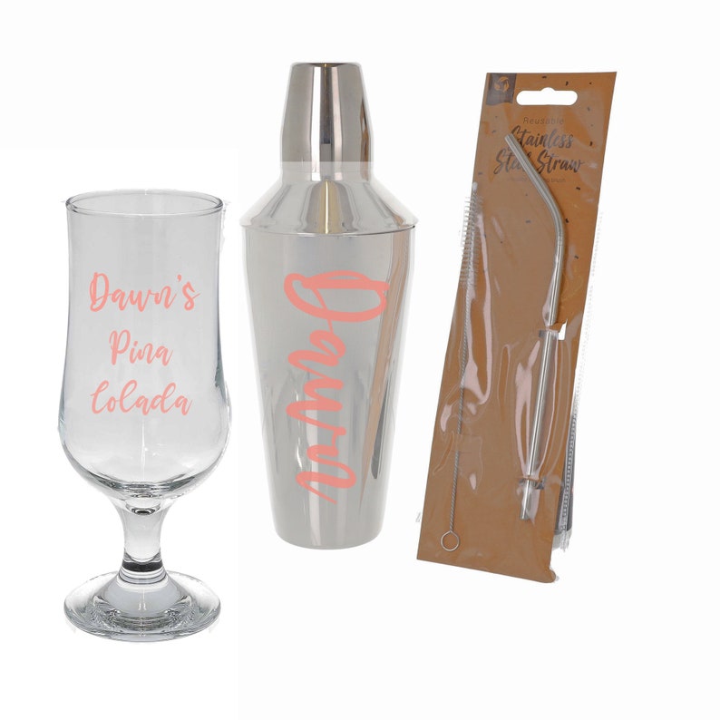 Puede incluir: Una coctelera de plata con el nombre "Dawn's" impreso en rosa en el lateral, un vaso transparente con el texto "Dawn's Pina Colada" impreso en rosa y una pajita de acero inoxidable con un cepillo de limpieza en un paquete de papel marr&oacute;n.