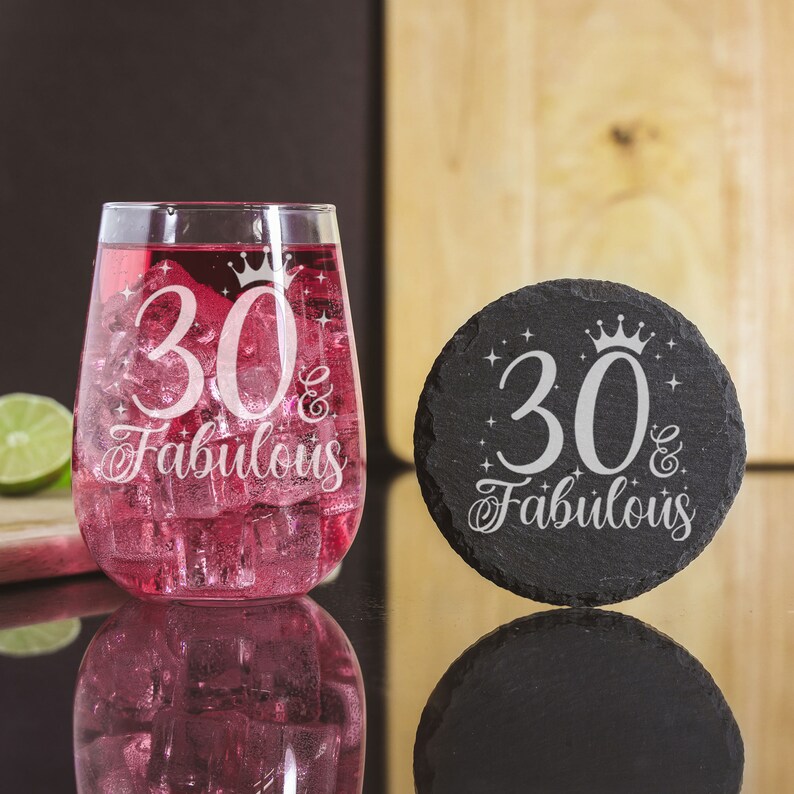May include: A black slate coaster with the text "30 & Fabulous" and a crown design. A clear glass with pink liquid and ice is next to the coaster.