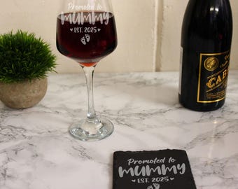 May include: A black slate coaster with the text "Promoted to Mummy Est. 2025" and a pair of footprints. A wine glass with the same text is in the background.