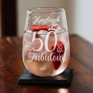 May include: A clear glass wine glass with the text "Kayleigh 50 & Fabulous" etched on the side. The glass is filled with a pink drink and ice.