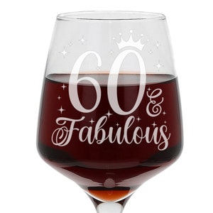 May include: A clear glass wine glass with a white design that says "60 & Fabulous" with a crown above the 60. The glass is filled with red wine.