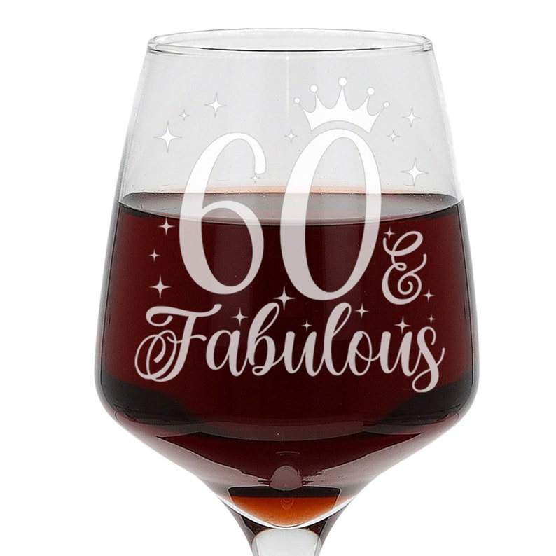 May include: A clear glass wine glass with a white design that says "60 & Fabulous" with a crown above the 60. The glass is filled with red wine.