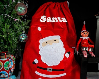 May include: Red Santa sack with a white felt Santa applique and the word "Santa" embroidered in white. A small elf doll is sitting on a black stool in the background.