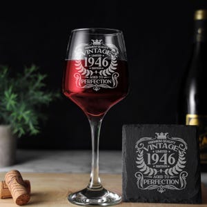 May include: A clear glass wine glass filled with red wine and a square black slate coaster, both etched with the text Premium Quality Vintage 1946 Limited Edition Aged to Perfection surrounded by a decorative laurel wreath and crown design.