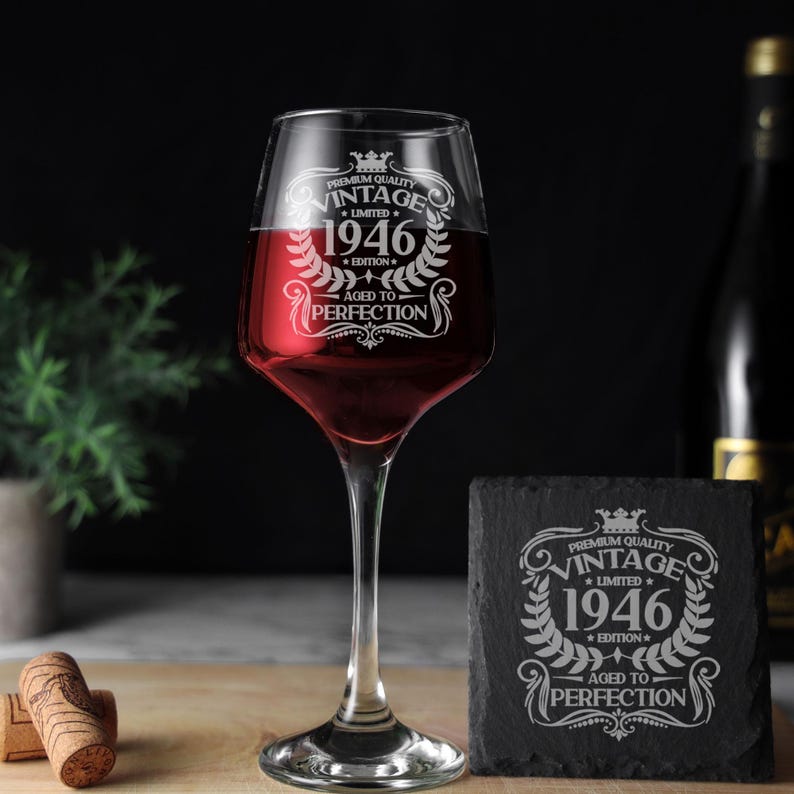 May include: A clear glass wine glass filled with red wine and a square black slate coaster, both etched with the text Premium Quality Vintage 1946 Limited Edition Aged to Perfection surrounded by a decorative laurel wreath and crown design.