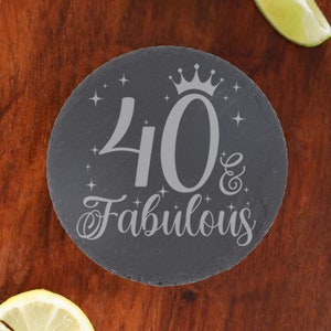 May include: A round black slate coaster with a silver inscription that reads "40 & Fabulous" with a crown above the number 40 and stars around the text.