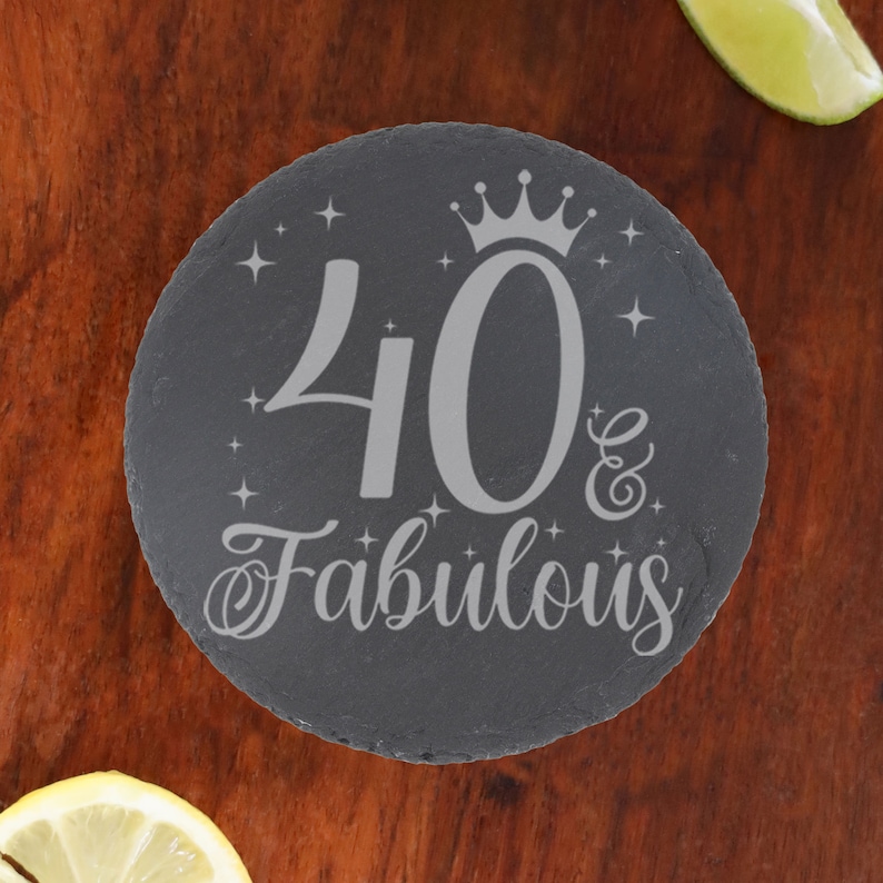 May include: A round black slate coaster with a silver inscription that reads "40 & Fabulous" with a crown above the number 40 and stars around the text.