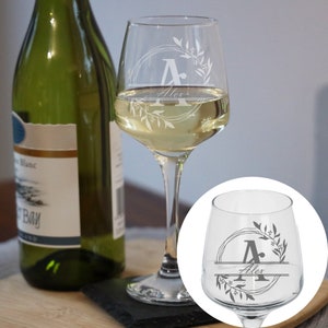 Puede incluir: Una copa de vino personalizada con un dise&ntilde;o de corona floral y la letra "A" en el centro. El nombre "Alex" est&aacute; grabado debajo de la letra. La copa est&aacute; llena de vino blanco.