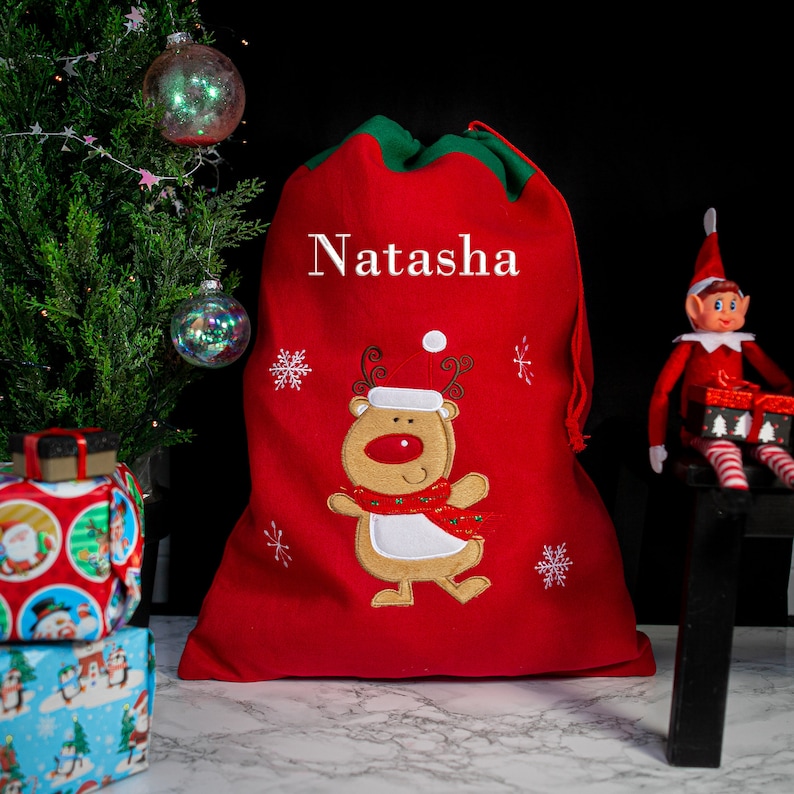 May include: Red personalized Santa sack with a reindeer design and the name Natasha embroidered on the front. The sack is decorated with snowflakes and has a green drawstring.