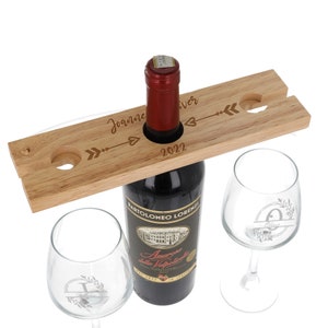 May include: Wooden wine bottle holder with two glass wine glasses. The holder has a cutout for a bottle and two slots for glasses. The holder is engraved with the names "Joanne" and "Oliver" and the year "2022".