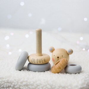 May include: A wooden stacking toy with a grey and natural wood finish. The toy features a bear with a pink nose and blue ears, and a base with the name "Oliver" engraved on it.